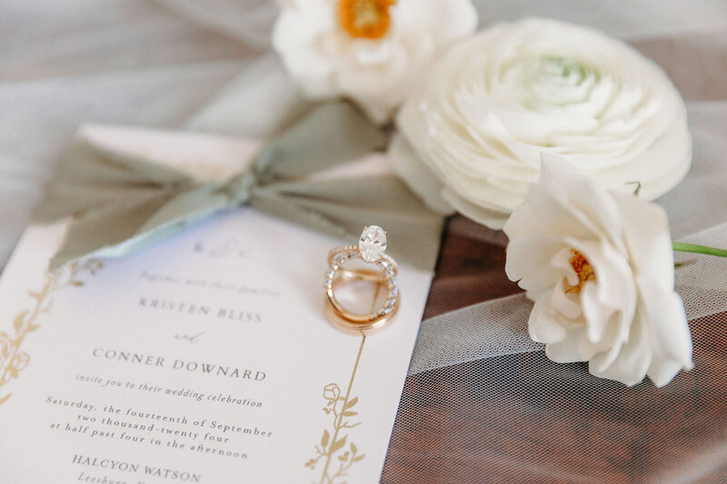 A wedding invitation rests on a sheer fabric adorned with white flowers and two rings, one featuring a large oval diamond. A gray ribbon embellishes the invitation, hinting at rustic wedding venue photos. The names Kristen Bliss and Conner Downard are elegantly displayed.