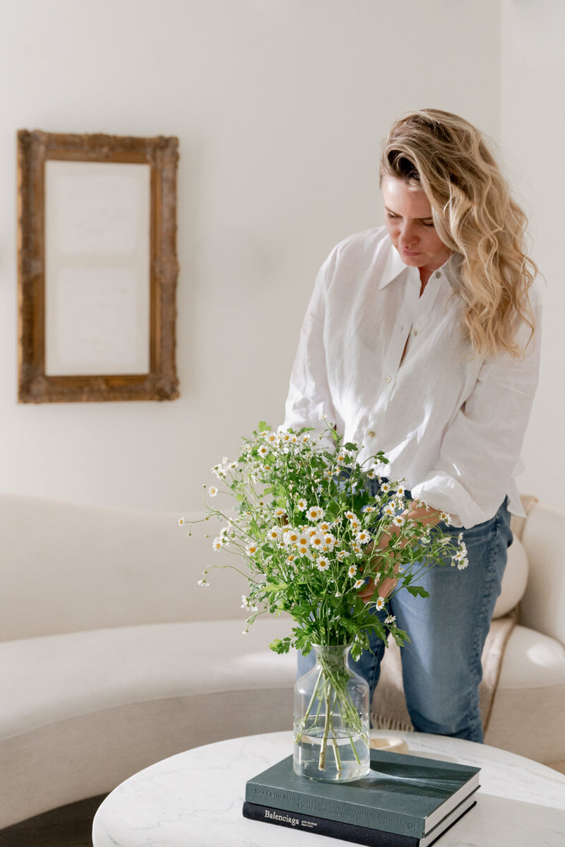 Blonde woman in a white shirt and jeans arranging flowers in a vase