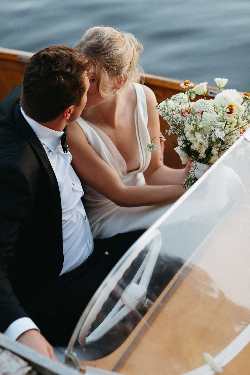 A couple in formal attire shares a kiss in a small boat, with the woman clutching a bouquet of flowers—a perfect moment captured by Toronto destination wedding photographer, Jennifer van Son at Muskoka Cottage.