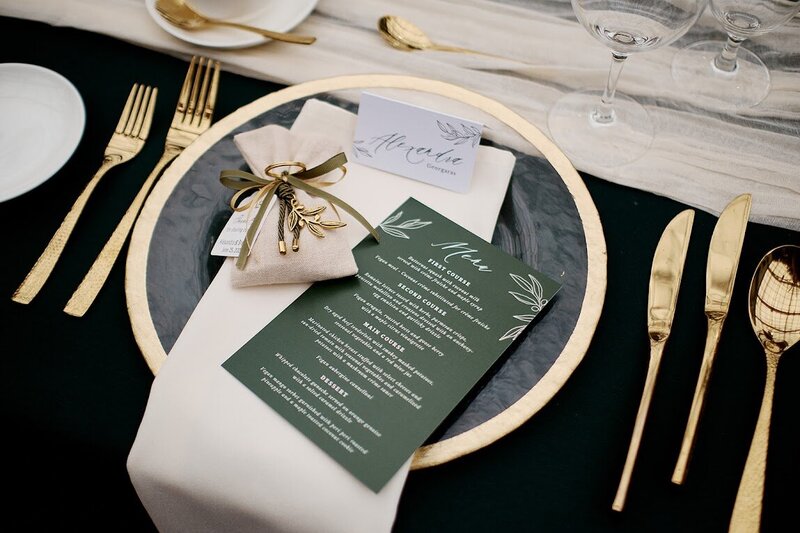 A minimalist forest green wedding menu sits on a gold-rimmed charger plate with gold cutlery beside it, on a black table linen for a wedding at Aquatopia  in Almonte, designed by Frid Events.