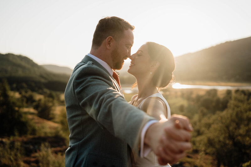 A couple stand with their chests together and arms extended. The link fingers and rub roses romantically. The wide photography shows golden hour on the Mar Lodge Estate and a large golden valley can be seen behind the couple. They wear wedding attire and the sun flickers between them.