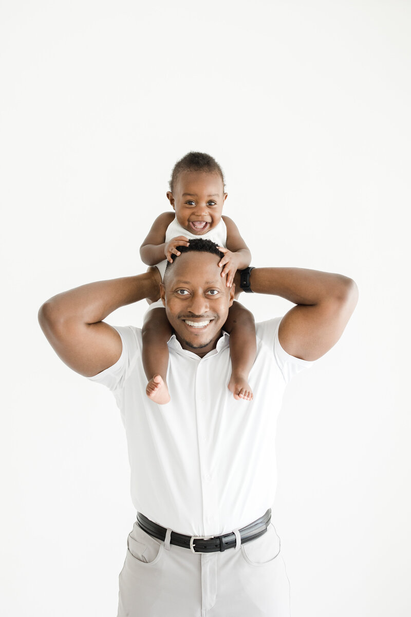Dad holds his one year old son on his shoulders as they both smile joyfully