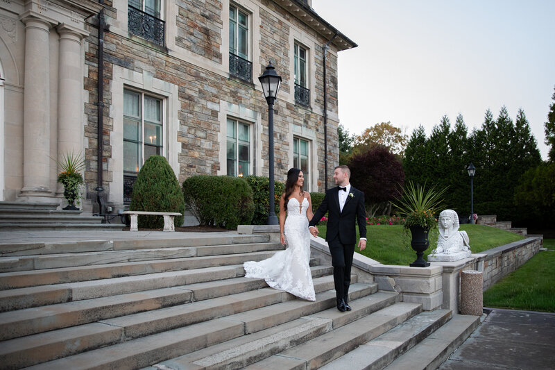 New England Wedding Photographer Pricing | Kelsey Sheehan Photography Timeless Rhode Island Weddings | A newlywed couple in elegant attire walks down stone steps, holding hands. They're beside a large stone building with lush greenery, exuding warmth and joy.