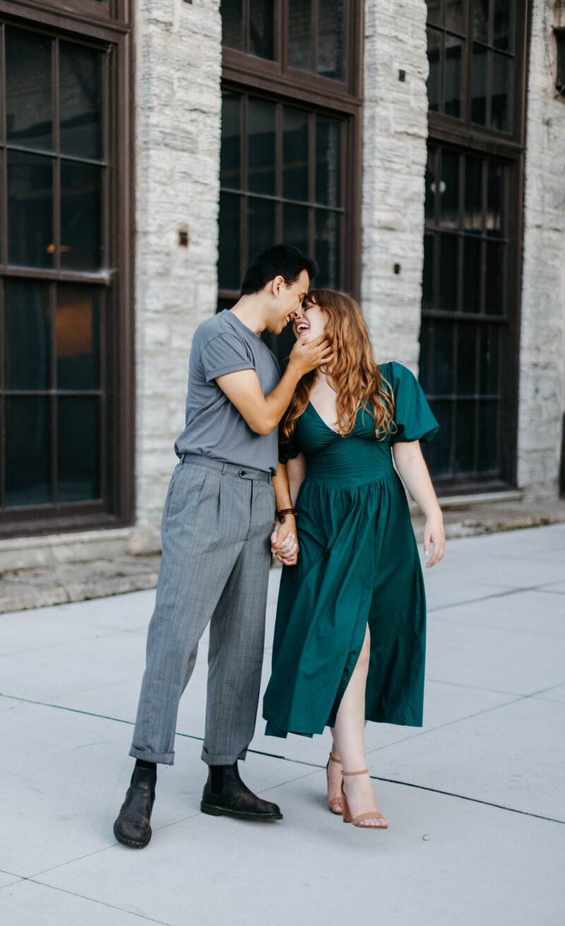 Couple posing for engagement photos outside in front of a building with tall windows