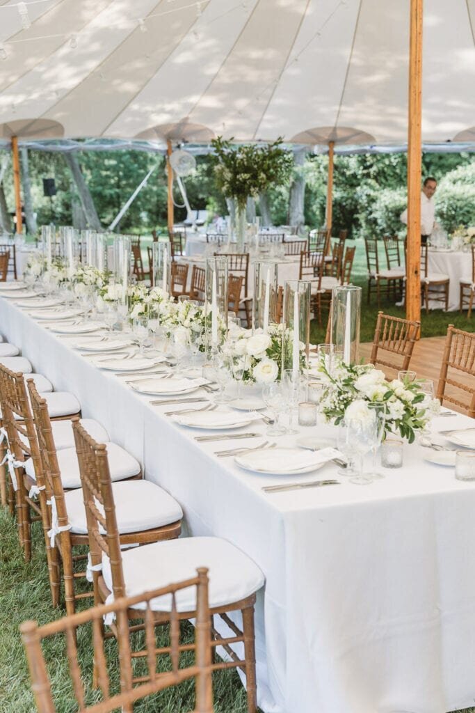 Sophisticated outdoor tented wedding reception with long tables, white floral centerpieces, and wooden chairs in a natural setting