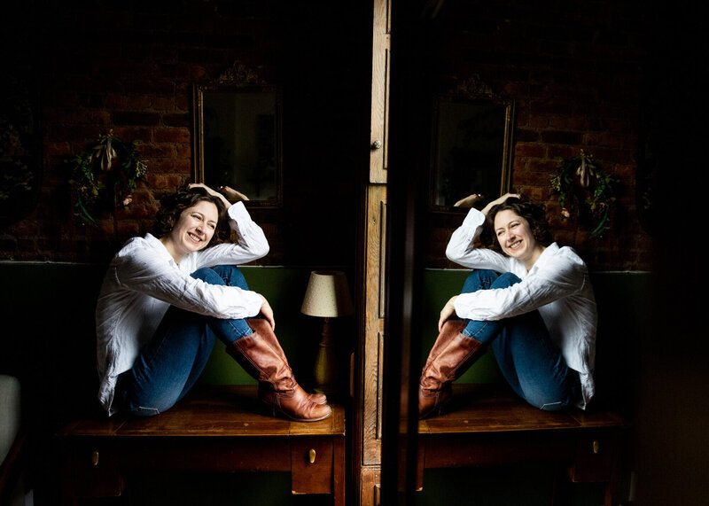 The photographer sitting on a table, smiling and looking out a window with her hand in her hair, reflected back in a mirror on the right so she appears to be sitting toe to toe with herself.