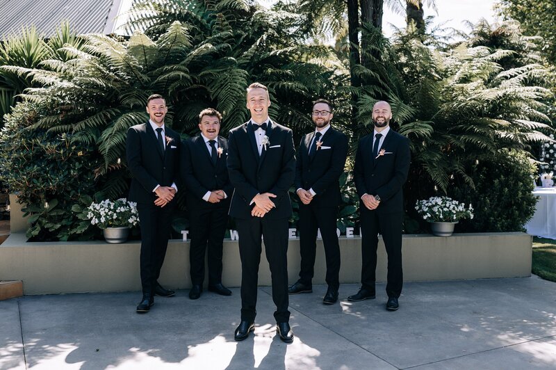 groomsmen in black tuxedos with black bow ties lined up in front of ferns in garden on christchurch wedding day