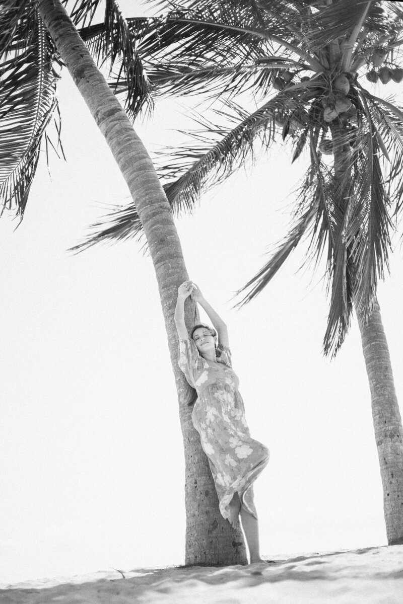 Model wearing swimsuit coverup leans against palm tree
