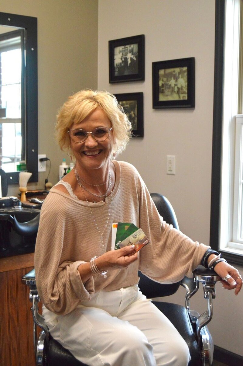 Martha Minier, the owner of 212 Salon, Spa, & Barbershop, sitting in a salon chair with a warm smile, holding color swatches, showcasing the welcoming and personalized service at her salon.