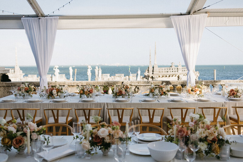 An outdoor wedding reception setup overlooking the water. The tablecloths are beige, the chairs are wooden with ivory cushions, and the centerpieces are a mix of peach, orange, and red florals