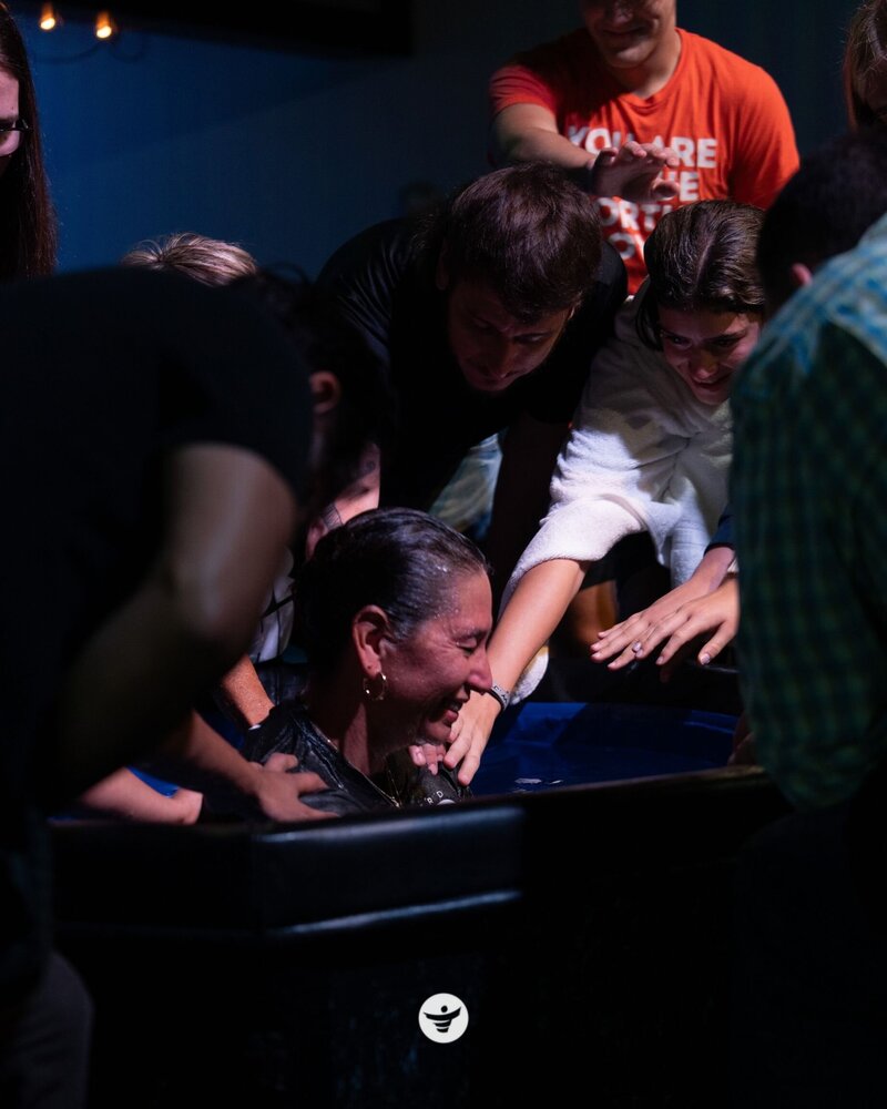 Woman in tub being baptized surrounded by smiling onlookers.