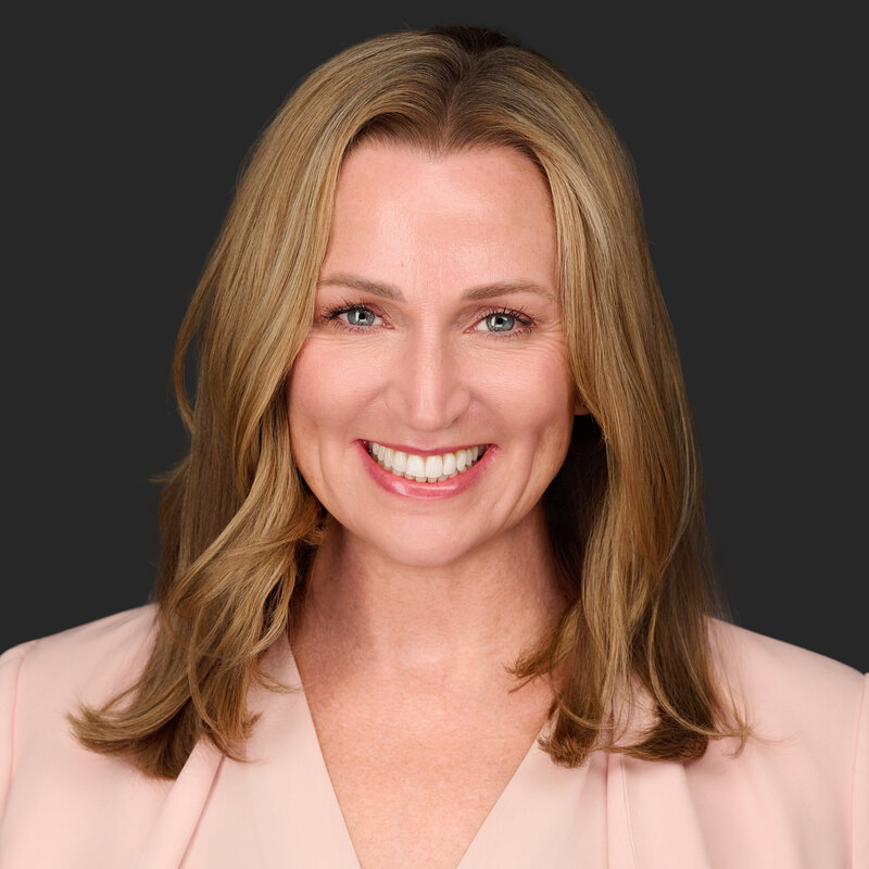 Jenn Cloutier, self-leadership coach, smiling warmly with shoulder-length blonde hair, wearing a light pink professional suit against a dark gray background.