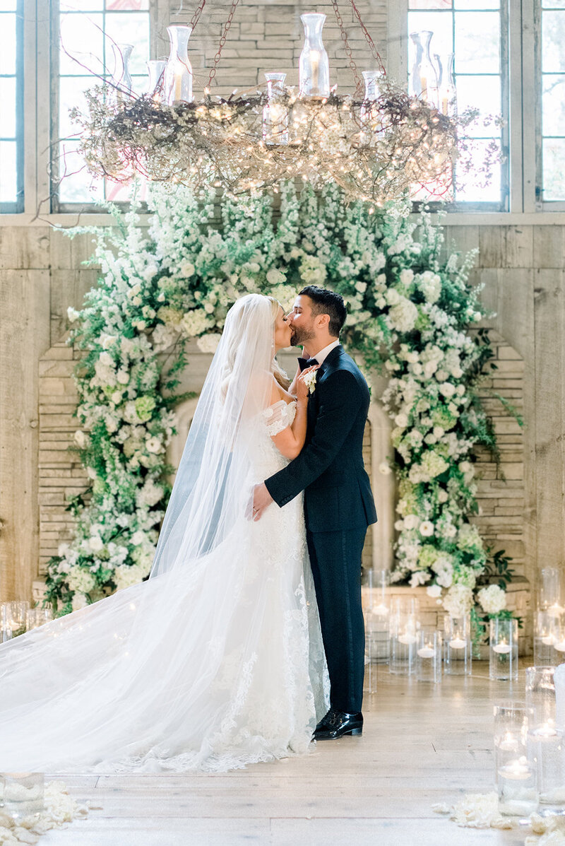 Tiffany Longeway Photography captures a romantic kiss between the bride and groom at The Estate Yountville in Napa Valley. The beautifully adorned floral arch and twinkling chandeliers create an enchanting and luxurious setting, perfect for couples seeking a sophisticated and memorable venue for their wedding celebration.