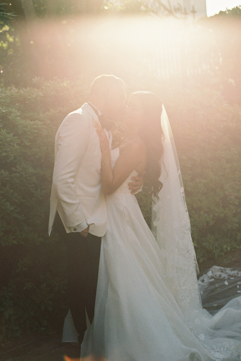 A bride leans on her groom's shoulder as they stand at their curtain altar with purple flowers