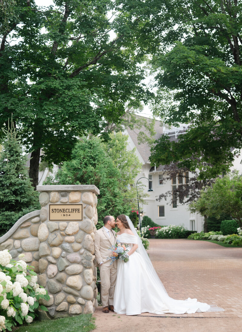 A couple stand in front of a old manor with many trees on a brick path at their  luxury destination  wedding on Mackinac Island at The Inn at Stonecliff.