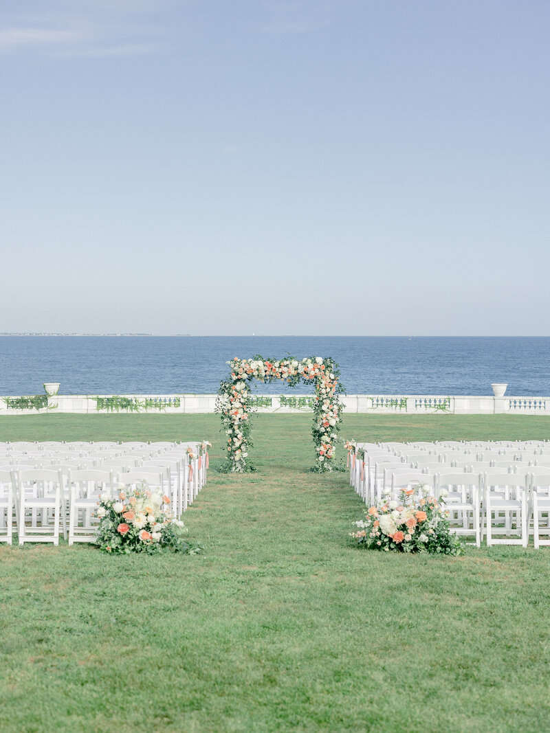 Outdoor wedding ceremony space with the ocean in the background and large floral arrangements on the ground along the aisle