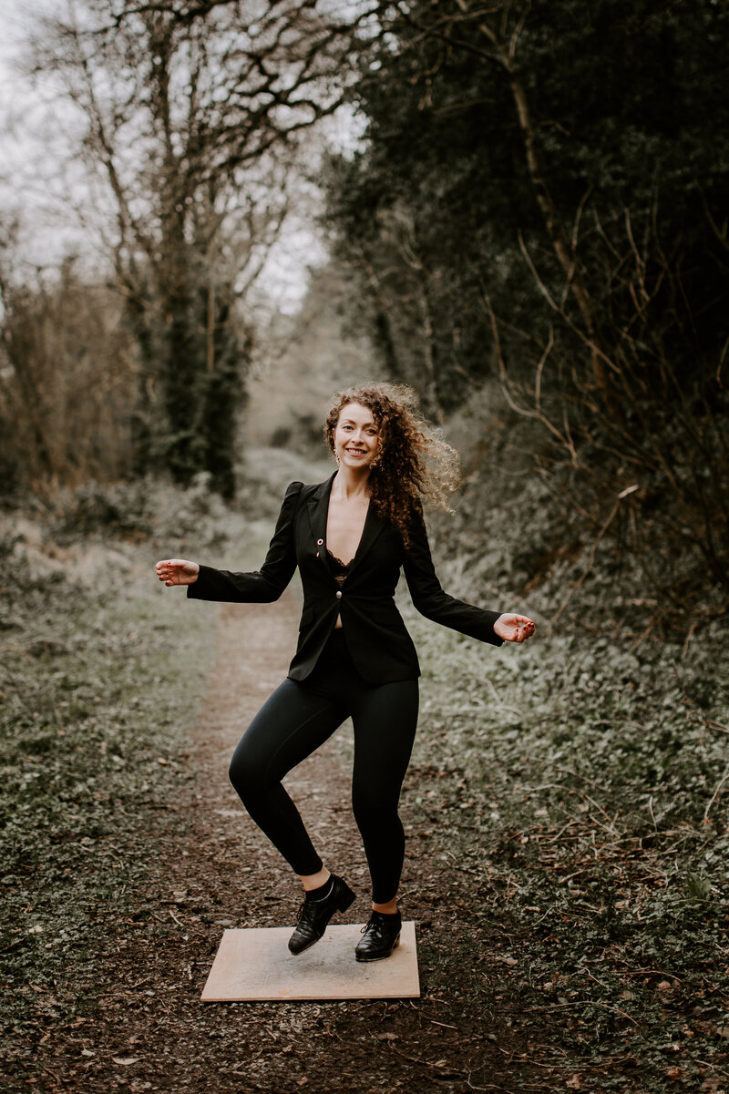 Seána Redmond smiling while performing sean nós dance outdoors in a woodland path, wearing a black blazer and trousers, representing Irish dance workshops and courses