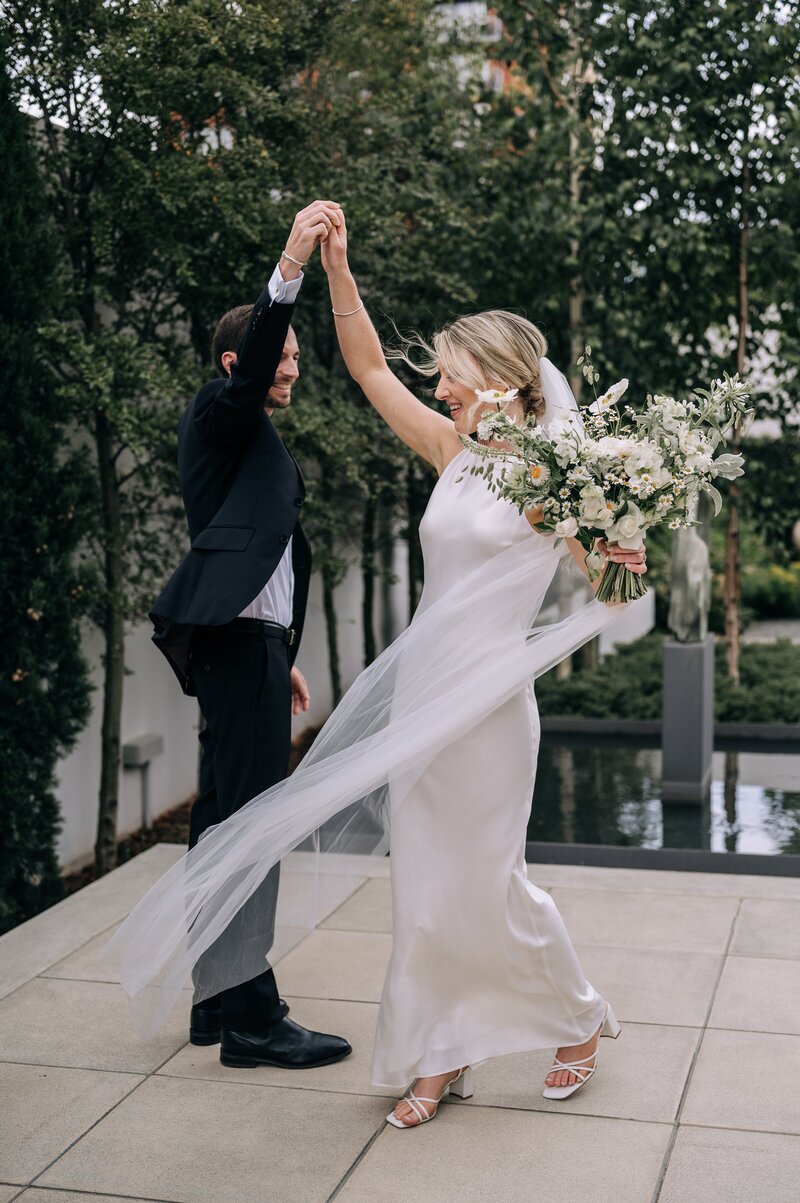 a groom in black suit and bowtie spins a bride with elegant ivory dress and loose white yellow and green bouquet