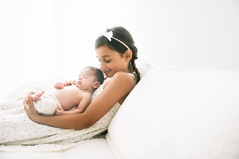 A young girl with braided hair and a white headband cradles a sleeping baby wrapped in a soft blanket. They are both on a light-colored bed with a bright, tranquil background.