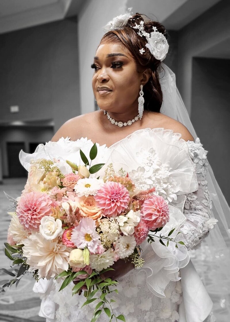 A bride in a luxurious wedding dress poses with a captivating expression. She wears an ornate floral headpiece and long, dangling earrings. Her makeup features dramatic eyes and glossy lips. She holds a large, stunning bouquet with pink dahlias, cream roses, and lush greenery, set against a soft-focus architectural interior background.