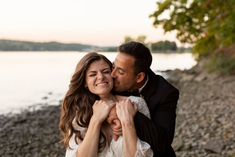 Groom hugs bride from behind while kissing her on the cheek.