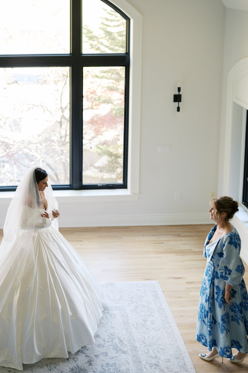 Bride in a voluminous gown and veil sharing a quiet moment with her mother, who is dressed in a floral blue dress, in a bright and airy room.