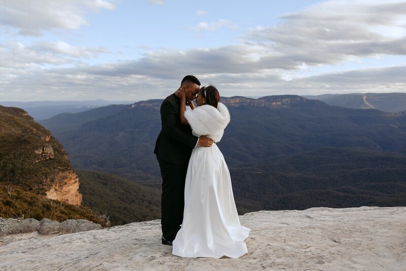 A bride in a fur jacket standing with her groom on the cliffs in the blue mountains
