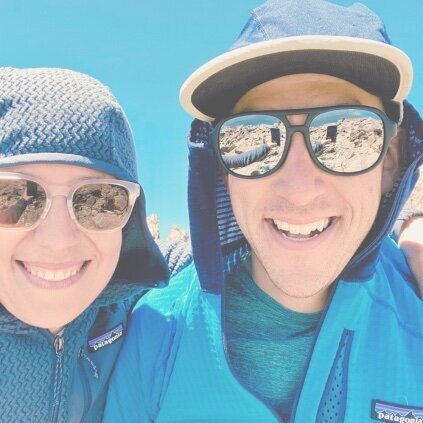 A woman and man wearing sunglasses smile directly at the viewer. The woman has her arm around the man and you can see her arm taking the photo reflected in the man's sunglasses. They are on the top of a rocky mountain with blue sky in the background.
