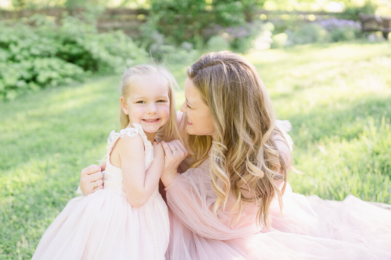 Sisters in matching pink dresses laying on a white blanket, looking at each other and smiling.