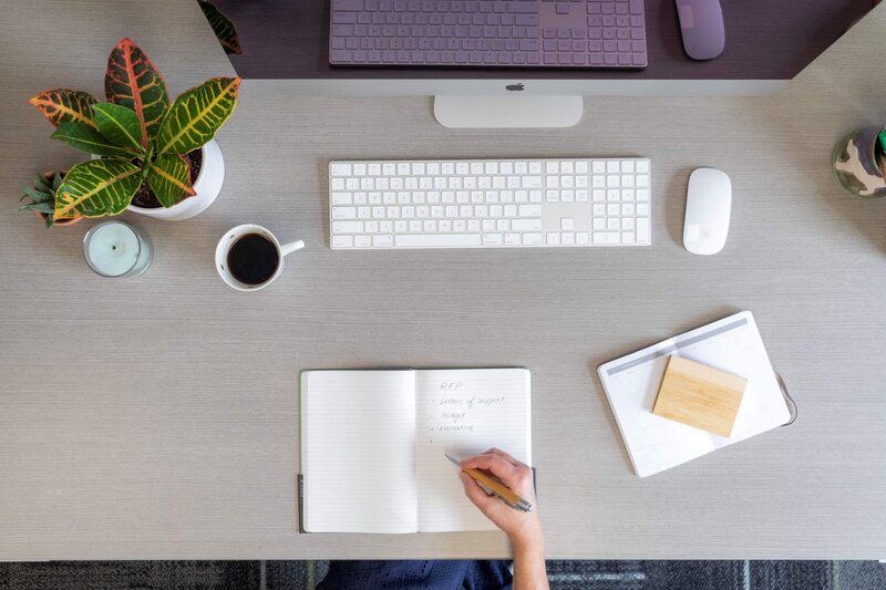 overhead view of desk with hand writing in a notebook, computer, coffee and plant on desk