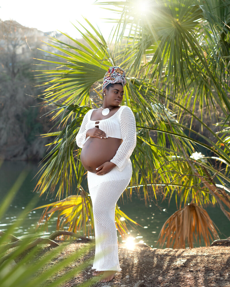 Pregnant woman in white crochet dress and colorful headwrap standing by Lake Travis during golden hour maternity session in Austin, Texas.