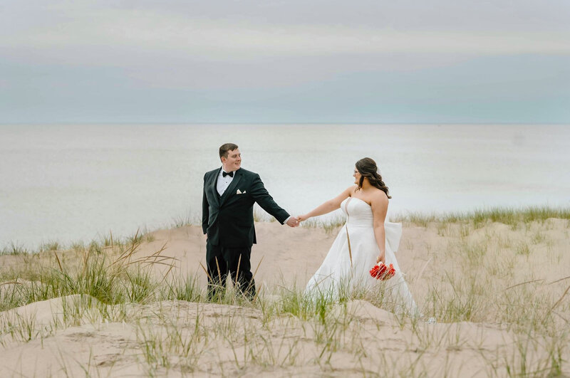 A bride and groom walk on the beach with their Michigan wedding photographer in Grand Haven at sunset