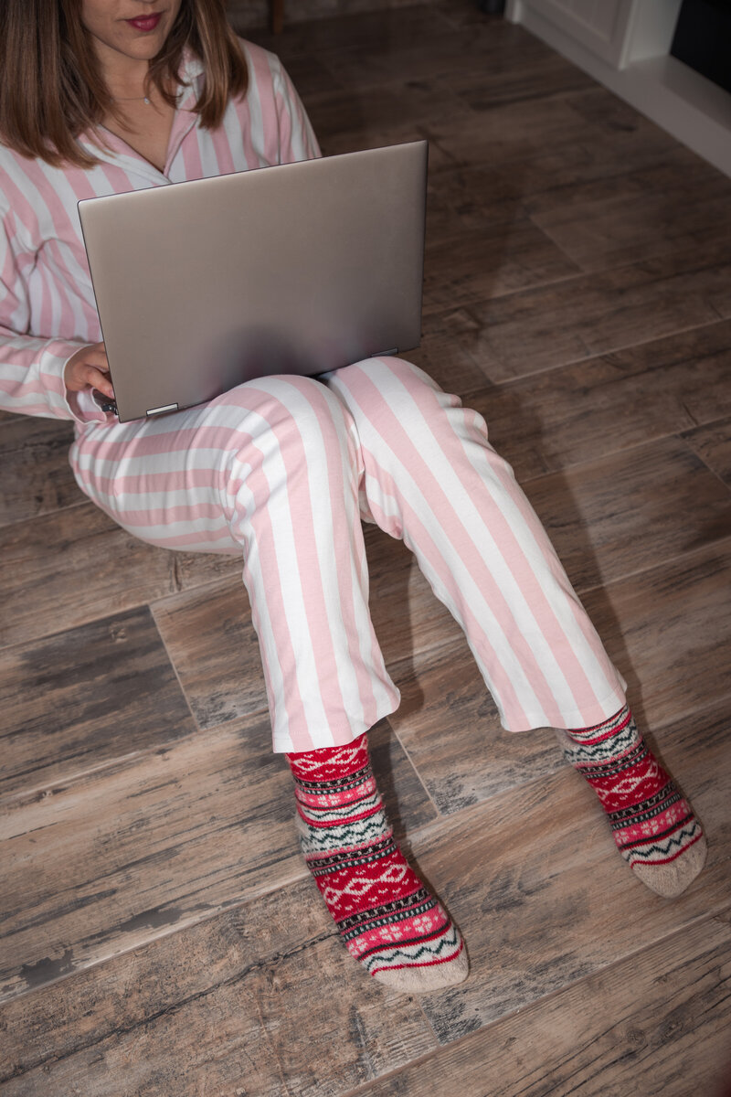 Girl sitting on the floor wearing pink and white stripped pajamas and colourful warm socks holding her laptop on her lap