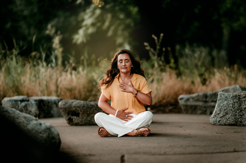 A picture of Danielle sitting doing breathwork outdoors hand on her heart and eyes closed