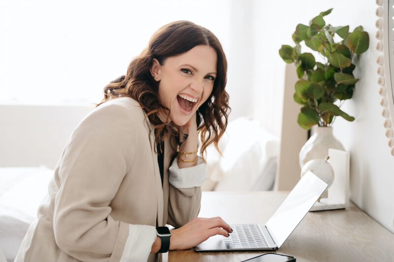 Woman laughing while using a laptop in a bright room during her photography mentorship.