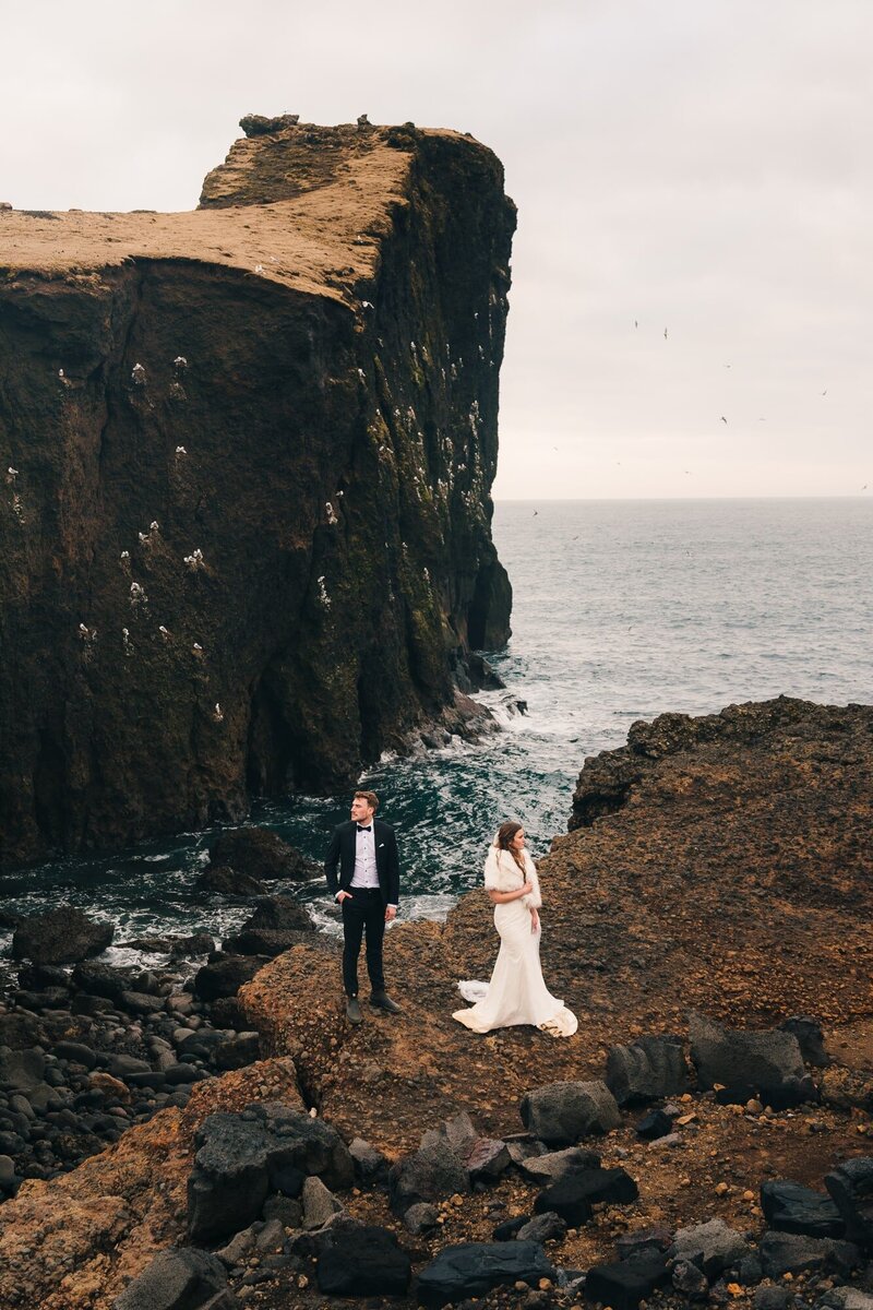 This couple shares a unique moment, gazing in opposite directions on jagged rocks during their elopement in Iceland, with the vast ocean as their breathtaking backdrop.