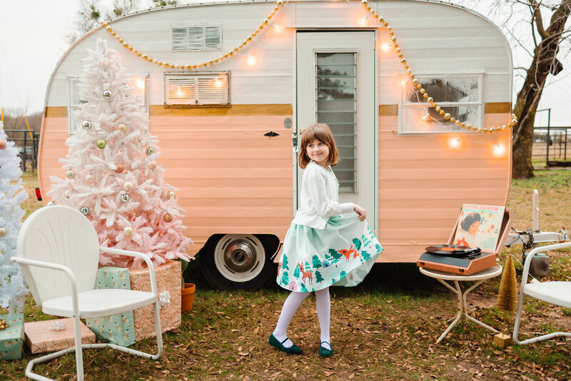 A girl poses in front of a pink mini car. She wears a blue dress and green ballet slippers.  Next to her, a pink Christmas tree and a white chair.