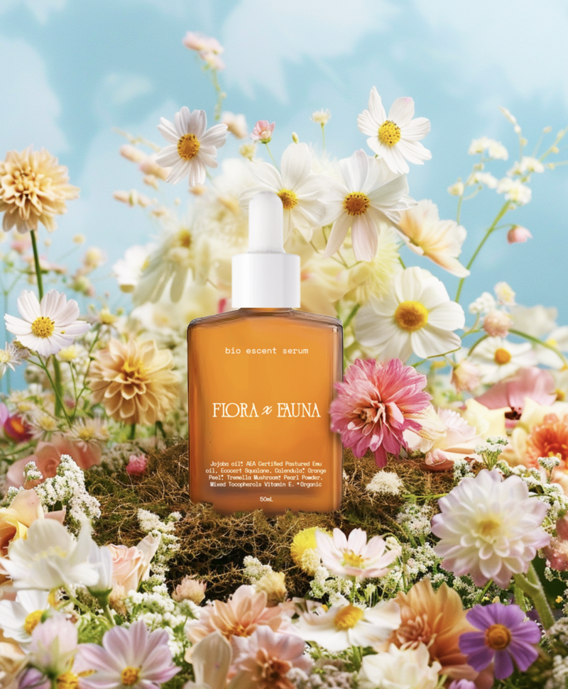 The Flora x Fauna Bio Accent Serum bottle is beautifully displayed amidst a lush arrangement of colorful flowers, set against a bright blue sky. The amber glass bottle contrasts with the soft pink, white, and yellow blooms, highlighting the serum’s natural and organic ingredients. The vibrant floral setting emphasizes the product’s connection to nature and its nourishing properties.