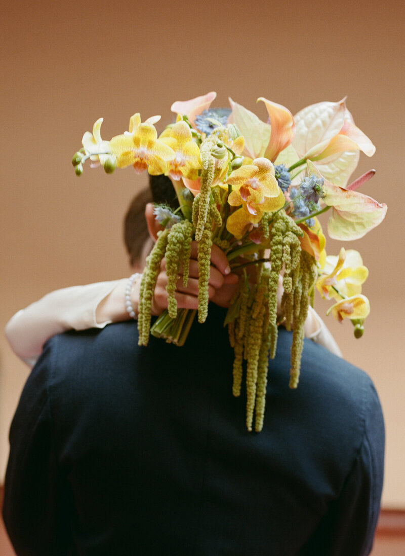 Bride’s bouquet of yellow flowers resting on groom’s shoulder, film photo.