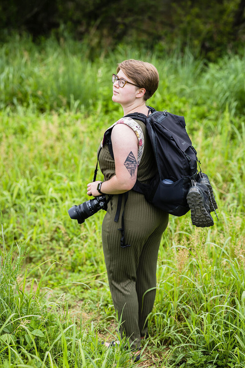 A Virginia wedding photographer wearing a hiking backpack  with hiking boots attached and camera on her shoulder looks over her shoulder with a slight smile in a field of grass.