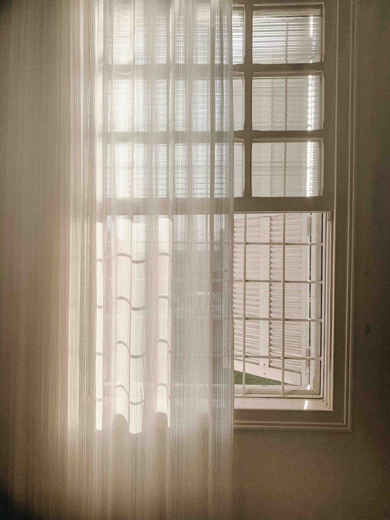 A cream colored window partially covered by a sheer ivory curtain