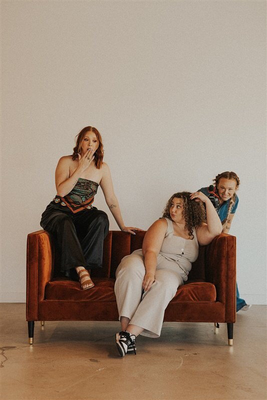 Three individuals strike playful poses on a burnt-orange couch. Steph, the person on the left, playfully covers their mouth; Andi, the person in the center, lounges with a thoughtful expression; and Lexi, the person on the right, leans over the backrest, smiling mischievously.