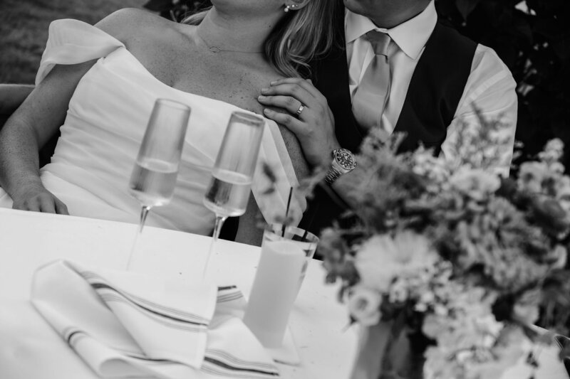 Black and white wedding reception table detail featuring champagne glasses, floral centerpiece, and the groom’s left hand with wedding ring resting gently on his bride’s shoulder.