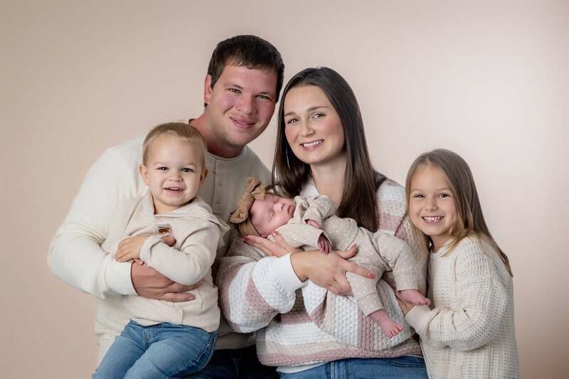 A happy family of 5 sits in a studio smiling with newborn sleeping in mom's arms