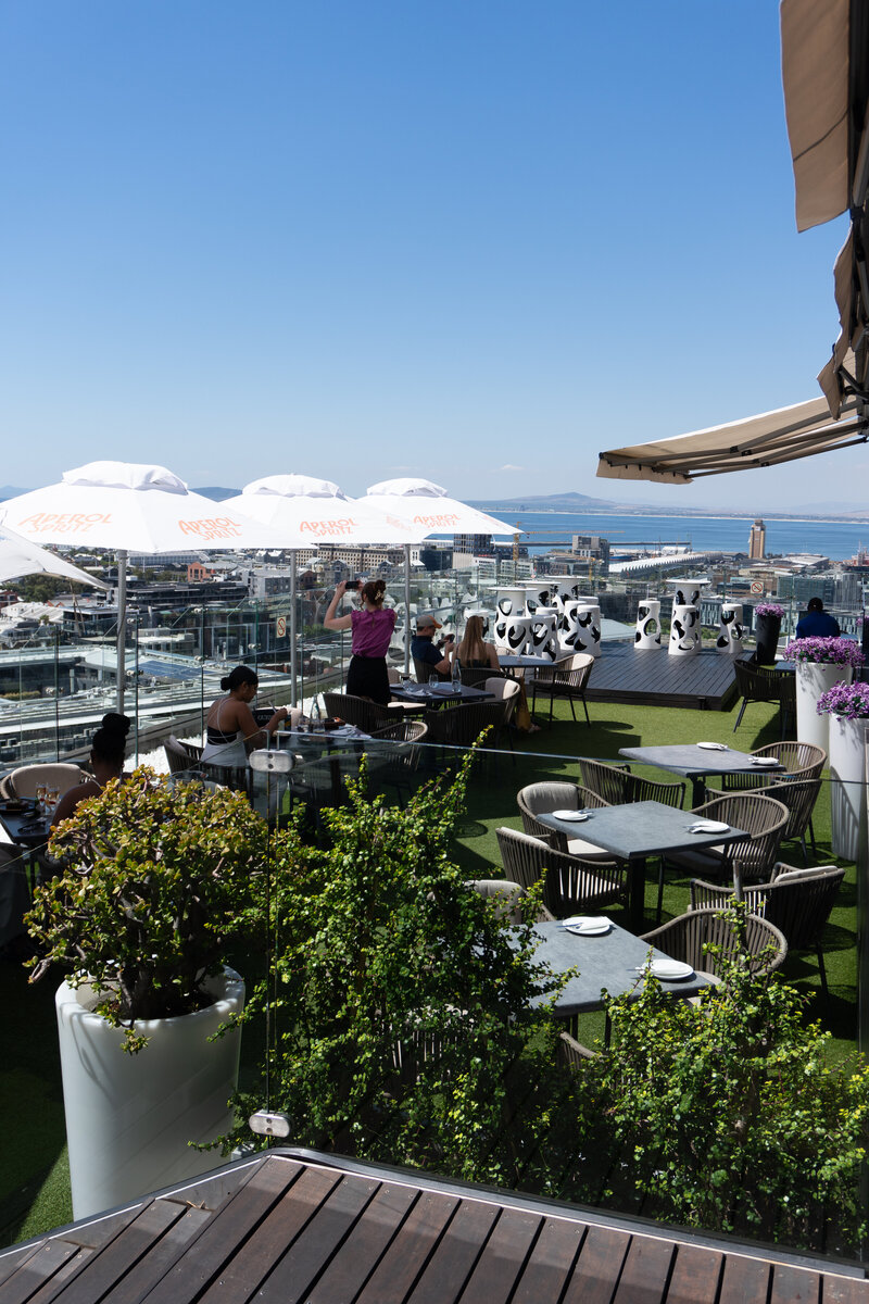 People dining on the rooftop of Utopia Cape Town Restaurant, overlooking a cityscape and the ocean.
