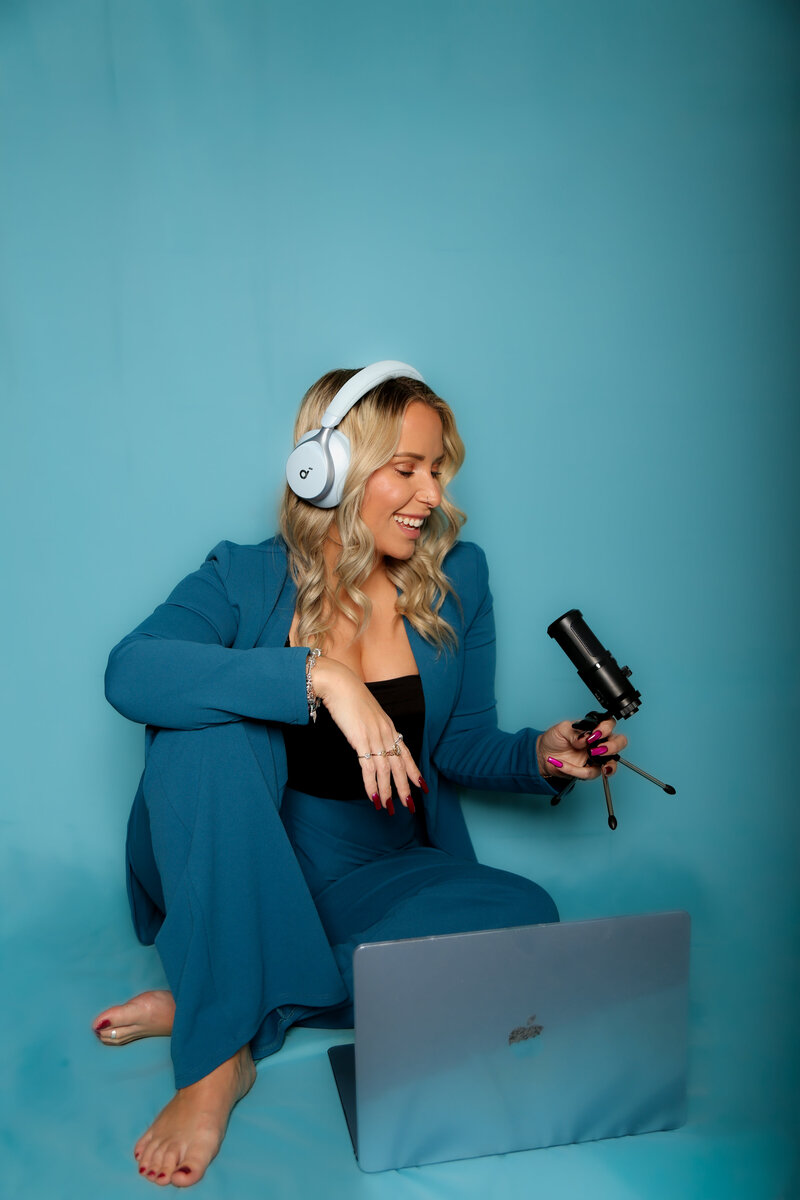 Robin wears a dark blue suit and sits on the floor with a laptop and a podcast microphone, recording content to help mothers in VA and beyond feel less alone in their motherhood journey