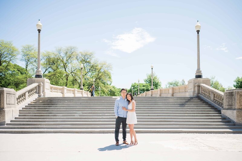 A couple poses affectionately at the base of a grand stone staircase in Chicago’s Lincoln Park, surrounded by historic lampposts and lush green trees under a bright blue sky.








