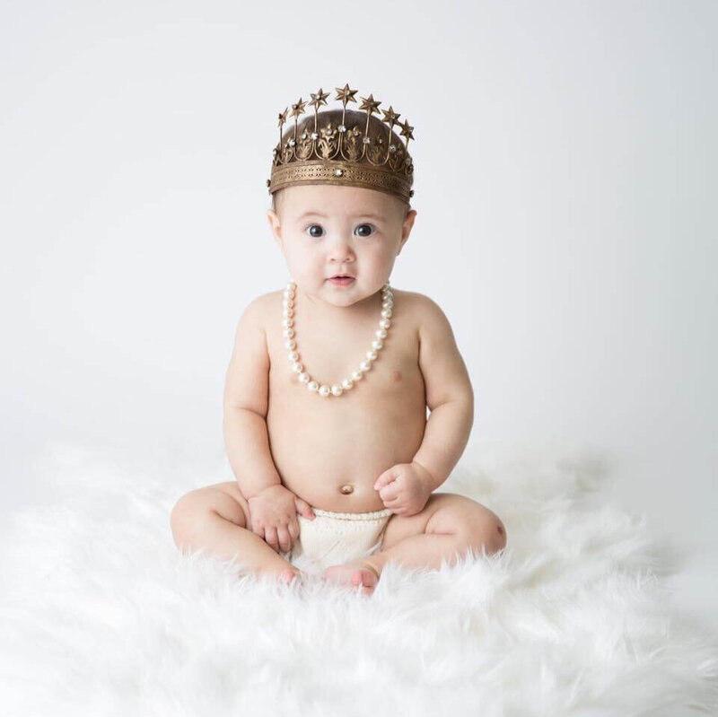 A baby wearing a decorative crown and pearl necklace sits on a fluffy white rug, looking curiously at the camera.