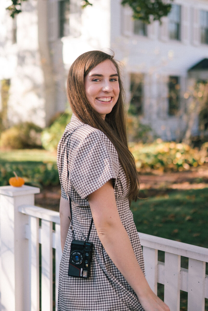 woman poses in front of white fence lined with pumpkins in downtown woodstock vermont in october