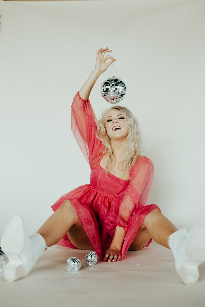 Sophie wearing a pink dress sitting on the floor holding a disco ball above her head